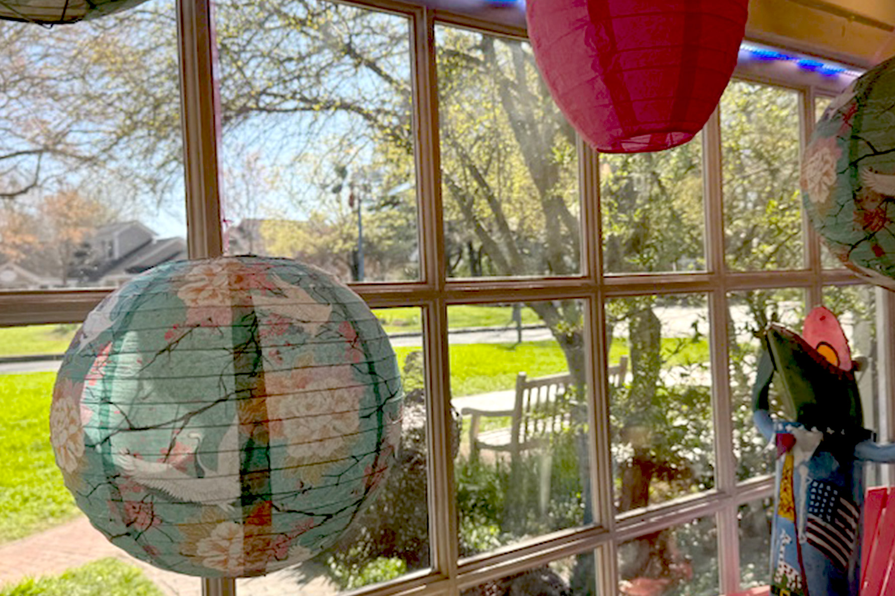 Colorful paper lanterns hang indoors, with large windows showing a sunny view of a park bench and trees outside. The bright daylight illuminates the vibrant colors of the lanterns against the serene greenery. Fearrington Village