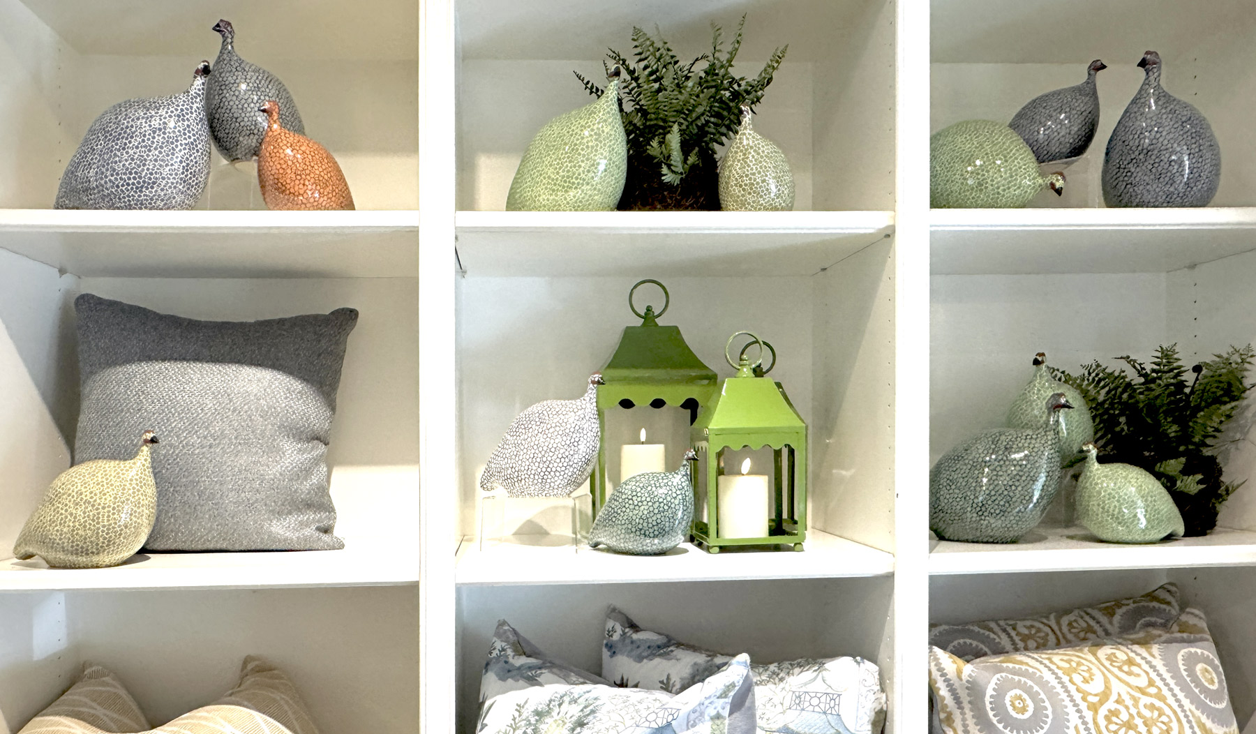 White shelves display decorative ceramic birds, green lanterns with candles, small potted ferns, gray and patterned pillows, and pastel-colored ornaments arranged neatly on each shelf. Fearrington Village