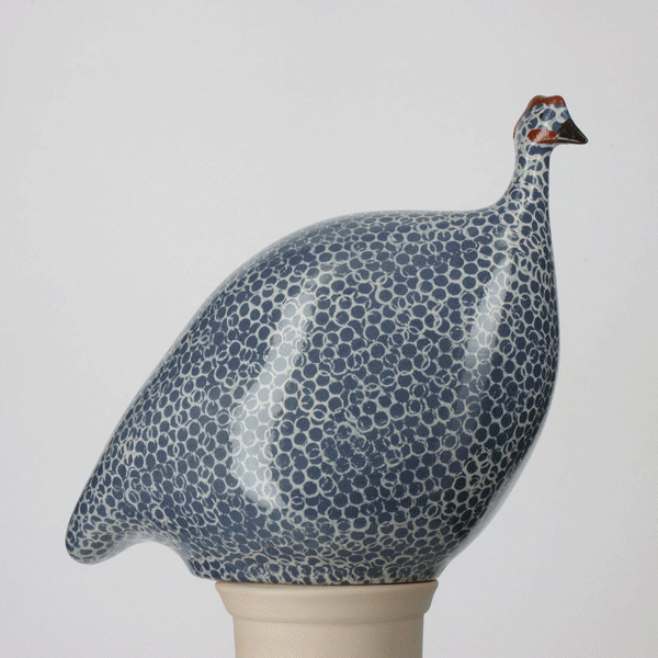 A ceramic sculpture of a guinea fowl with a round, speckled blue and white body, a small head, and a short neck, displayed against a plain light background. Fearrington Village