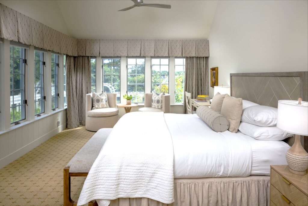 A spacious, well-lit bedroom features a large bed with white linens and pillows, a wooden bench at the foot, two beige chairs by the windows, a small round table with a plant, and a lamp on the nightstand. The room has many windows and neutral tones. Fearrington Village