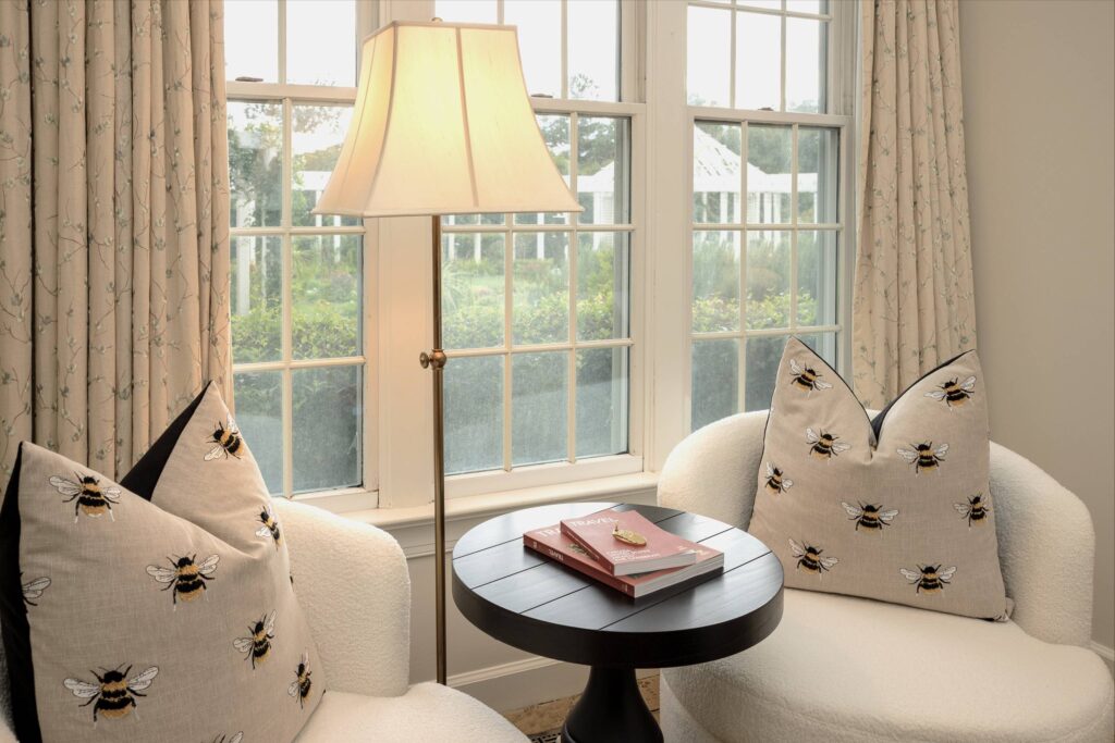 Cozy seating area with two white chairs and a round black table in between. Bee-patterned pillows rest on the chairs. A lamp with a beige shade stands beside tall windows with cream curtains, overlooking greenery outside. A book and decorative item are on the table. Fearrington Village