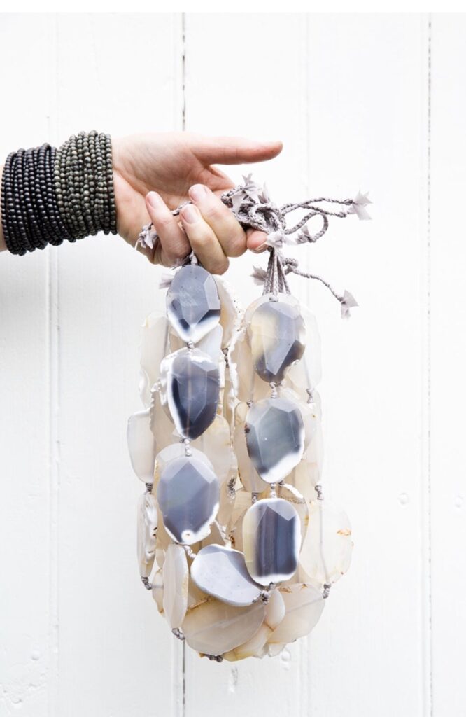 A hand wearing black beaded bracelets holds a string of large, glossy, oval stones in shades of white and gray. The stones are tied together with a knotted gray cord, standing out against a white wooden background. Fearrington Village