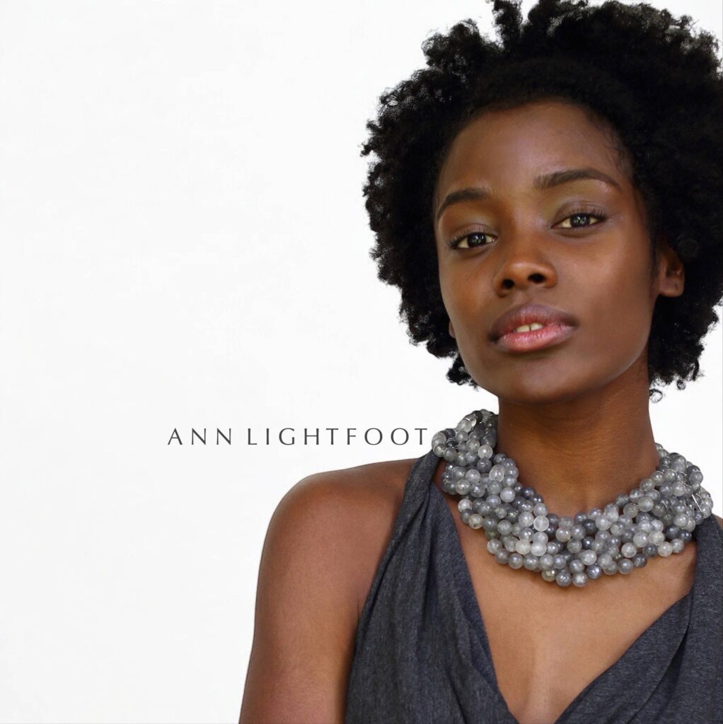 A person with curly hair models a chunky, gray beaded necklace against a plain white background. They wear a gray sleeveless top and the name "Ann Lightfoot" appears in the middle of the image. Fearrington Village
