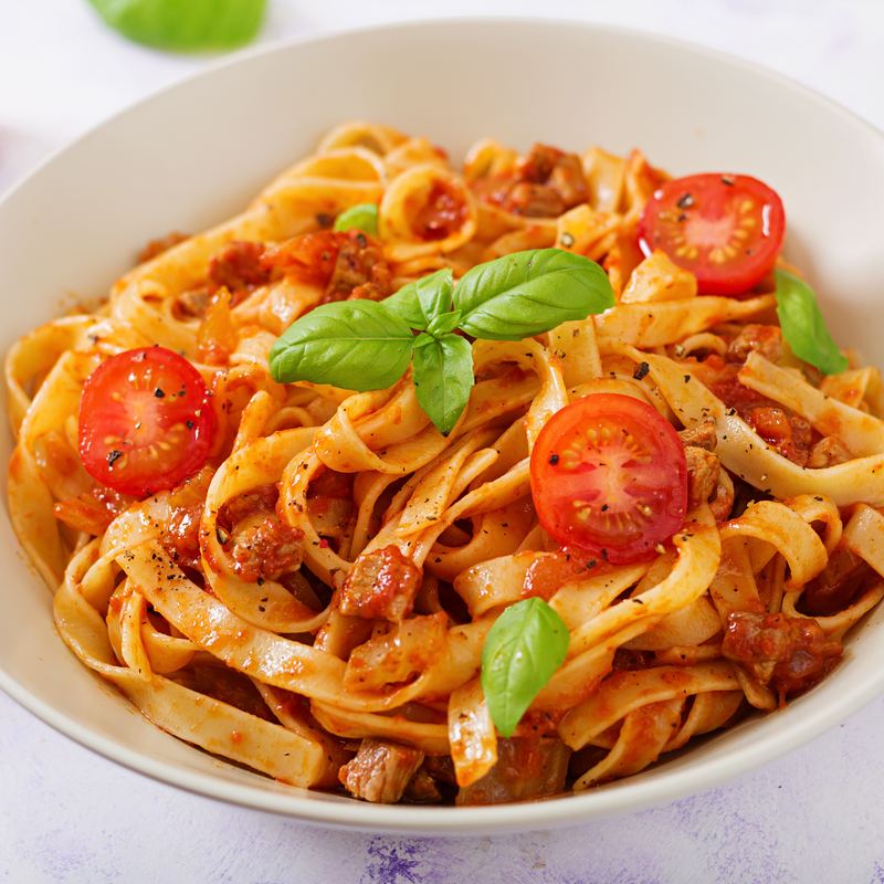 A bowl of fettuccine pasta in a tomato-based sauce with chunks of meat, garnished with halved cherry tomatoes and fresh basil leaves. The dish is sprinkled with black pepper, adding a savory touch. Fearrington Village