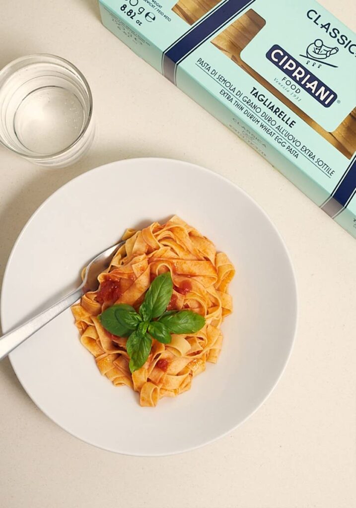 A plate of tagliatelle pasta with tomato sauce garnished with fresh basil sits on a white table. A fork rests on the plate's edge. A glass of water and a box of Cipriani tagliatelle pasta are placed nearby. Fearrington Village