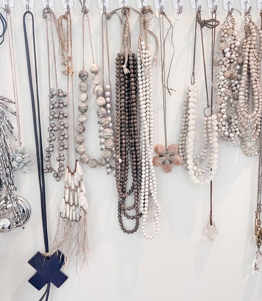 A collection of various beaded necklaces hanging on hooks against a white wall. The necklaces are made of white, gray, and brown beads, with designs including tassels and a pendant in the shape of a plus sign. Fearrington Village