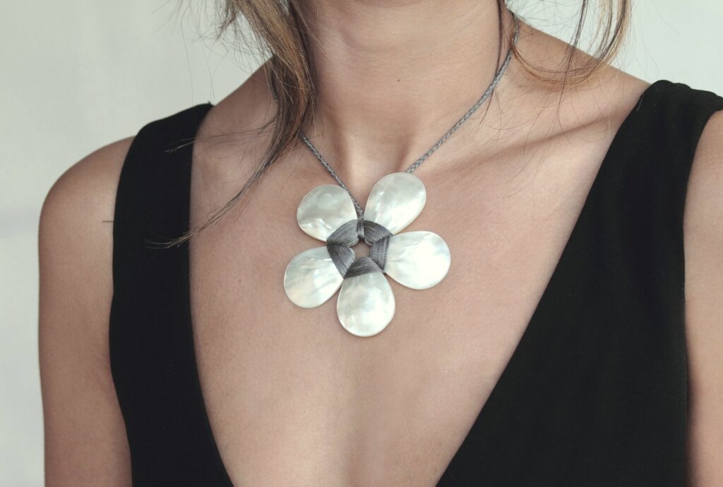 A woman wears a large flower-shaped necklace with silver and pearl-like petals on a thin chain. She is dressed in a black V-neck top. The background is neutral, highlighting the jewelry. Fearrington Village