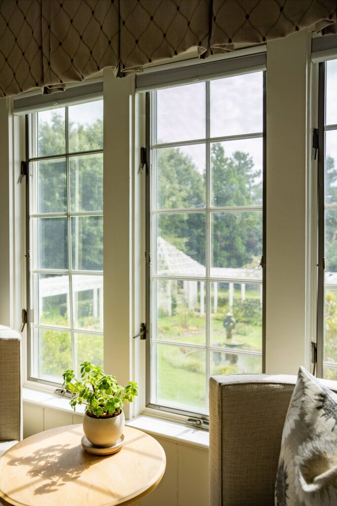 A sunlit room with two large windows overlooking a green garden. A small round table sits between two chairs, holding a potted plant. The outside view includes trees and a pergola under a blue sky. Fearrington Village