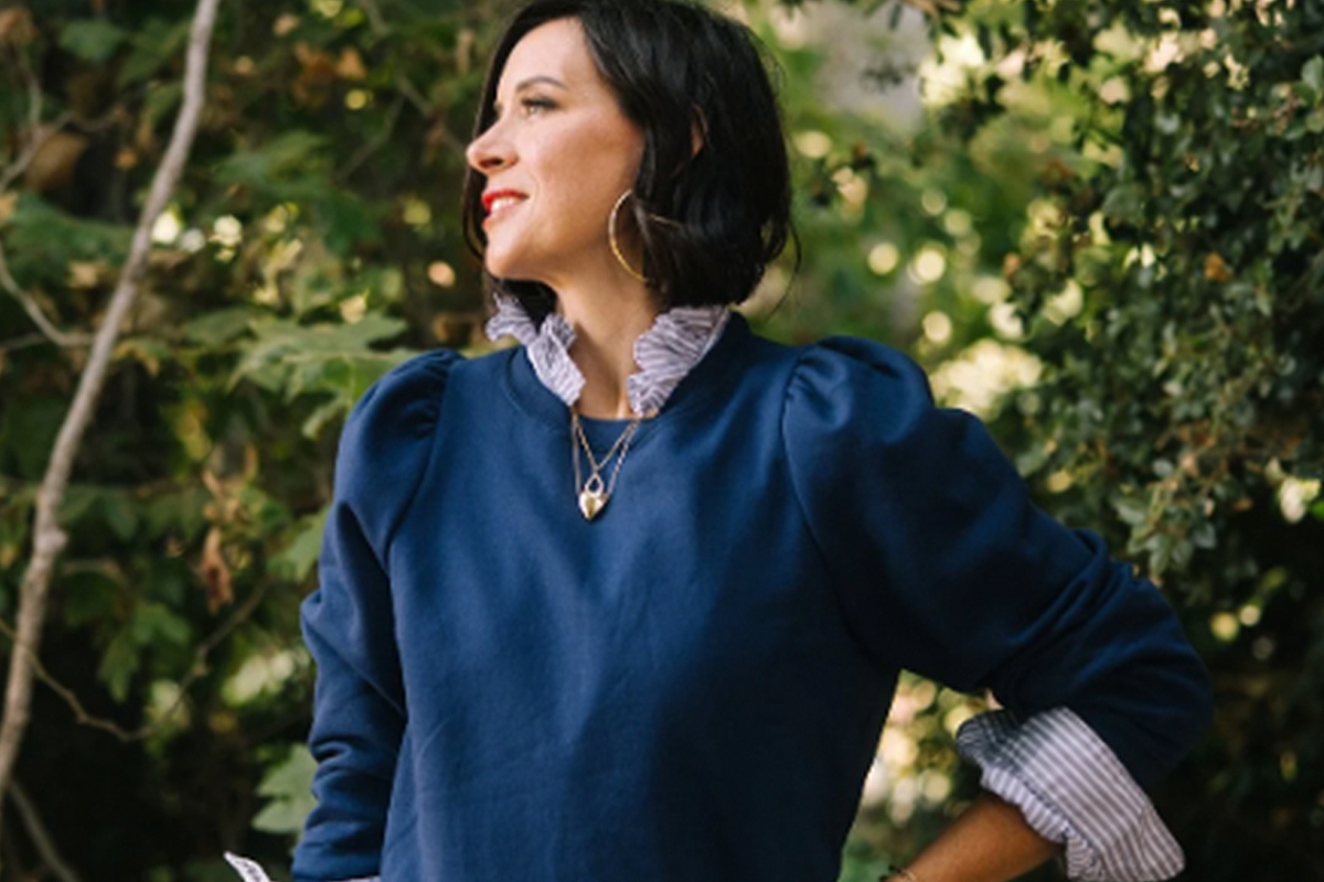 A person with dark hair wearing a blue blouse and gold jewelry is standing outdoors. The blouse has ruffled cuffs and the necklace has two pendants. The background is filled with green foliage. The person is smiling and looking to the side. Fearrington Village