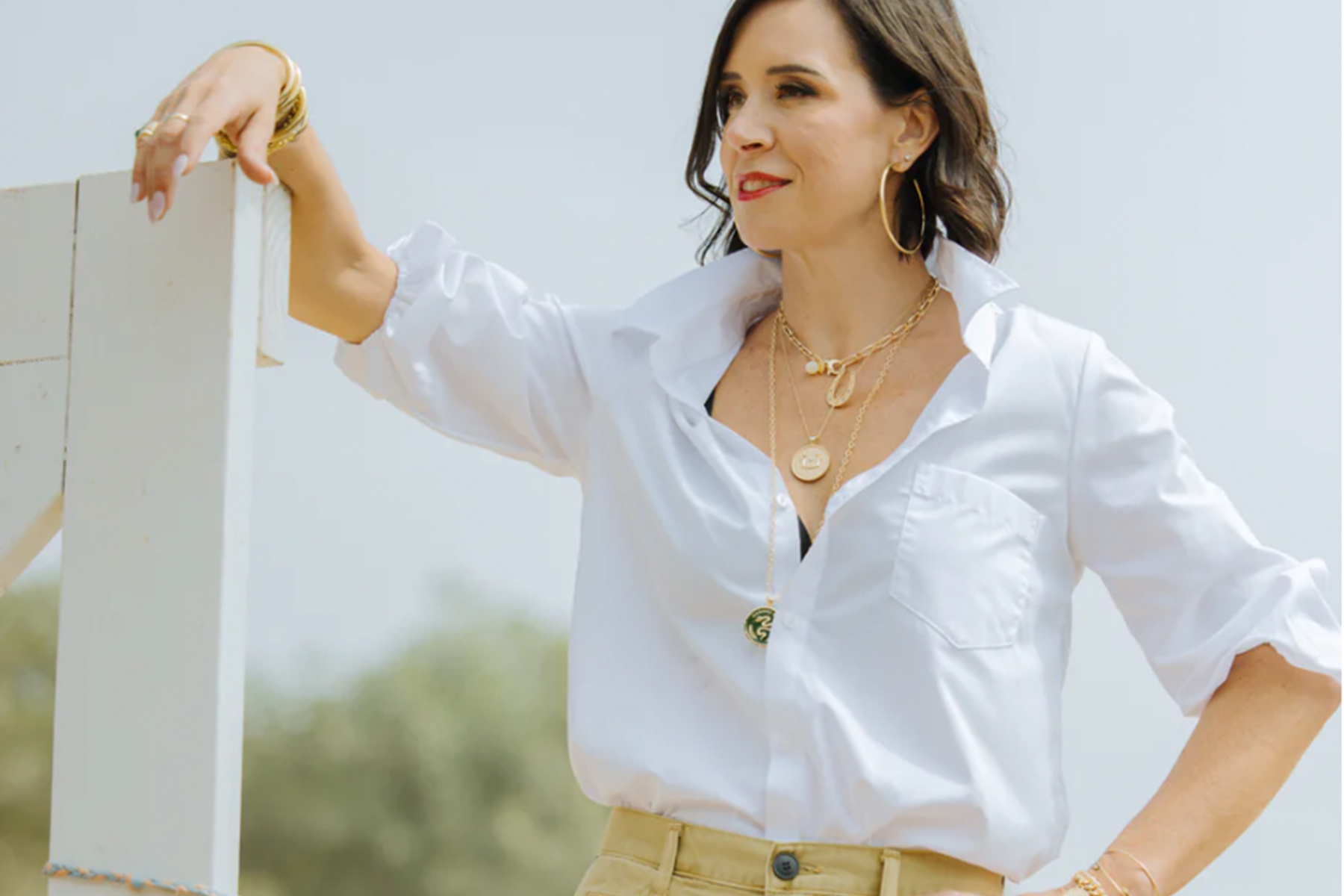 A woman with dark hair, wearing a white blouse and beige pants, stands confidently. She is accessorized with hoop earrings and layered necklaces. Her expression is relaxed with a slight smile, and the background is a clear sky and blurred greenery. Fearrington Village