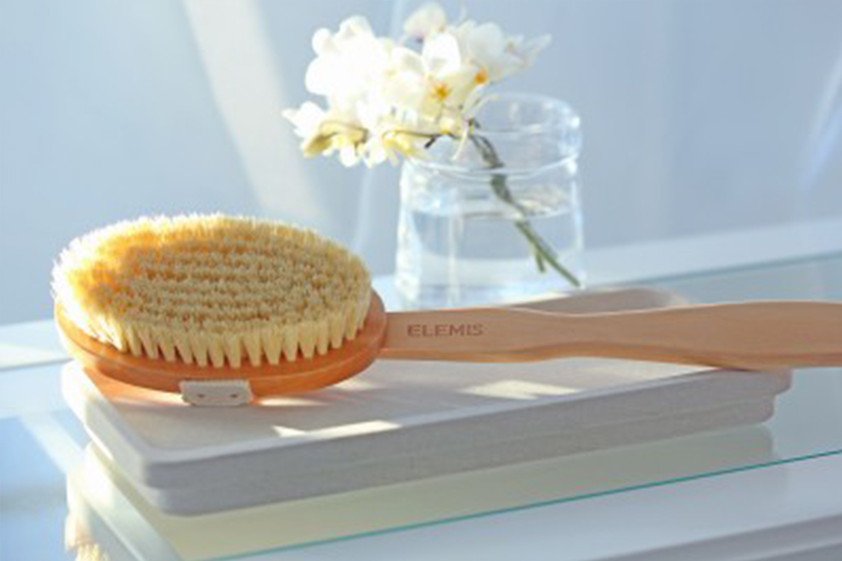 A wooden-handled body brush with soft bristles rests on a white folded towel. In the background, there's a glass with water and white flowers on a glass surface, all bathed in soft natural light. Fearrington Village