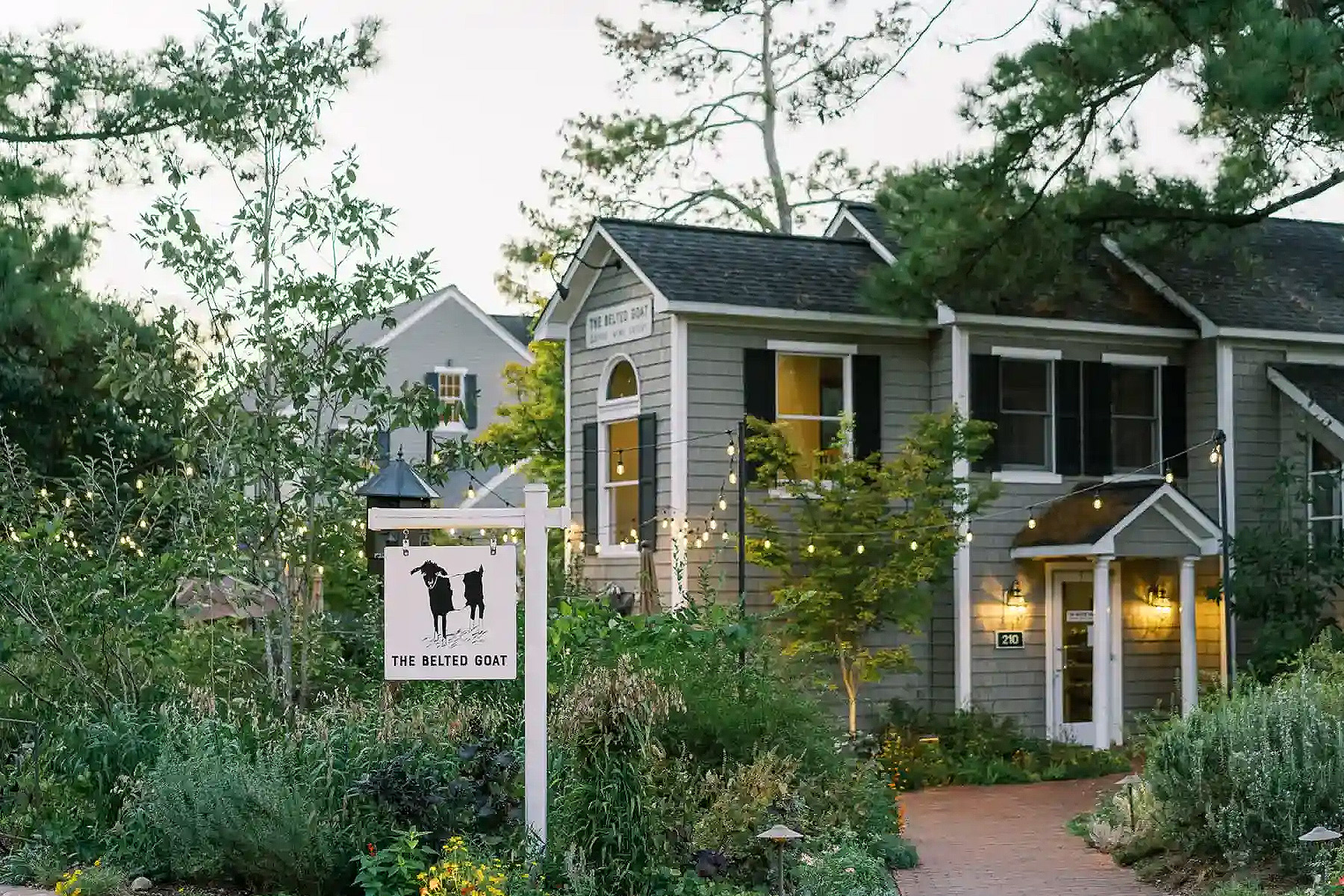 A quaint gray building with white trim, surrounded by lush greenery and string lights. A sign reads "The Belltol Goat," featuring a silhouette of a goat. The setting is peaceful, with a brick pathway leading to the entrance. Fearrington Village