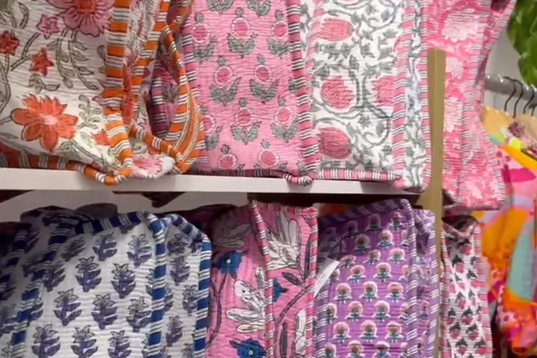 Folded, colorful quilted blankets are neatly stacked on shelves. The designs include floral and geometric patterns in various colors like pink, blue, orange, and white. The texture appears soft and cozy, inviting a sense of warmth. Fearrington Village