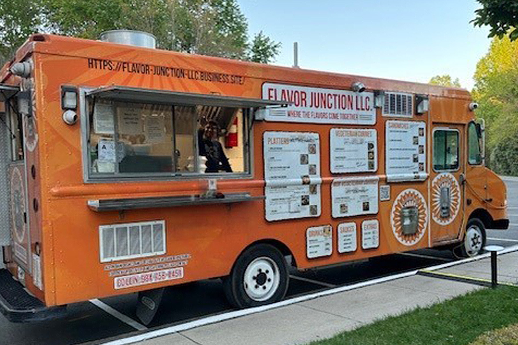 An orange food truck labeled "Flavor Junction LLC" is parked by a curb, with its serving window open. Menus and signs cover the side, and trees are visible in the background. Fearrington Village
