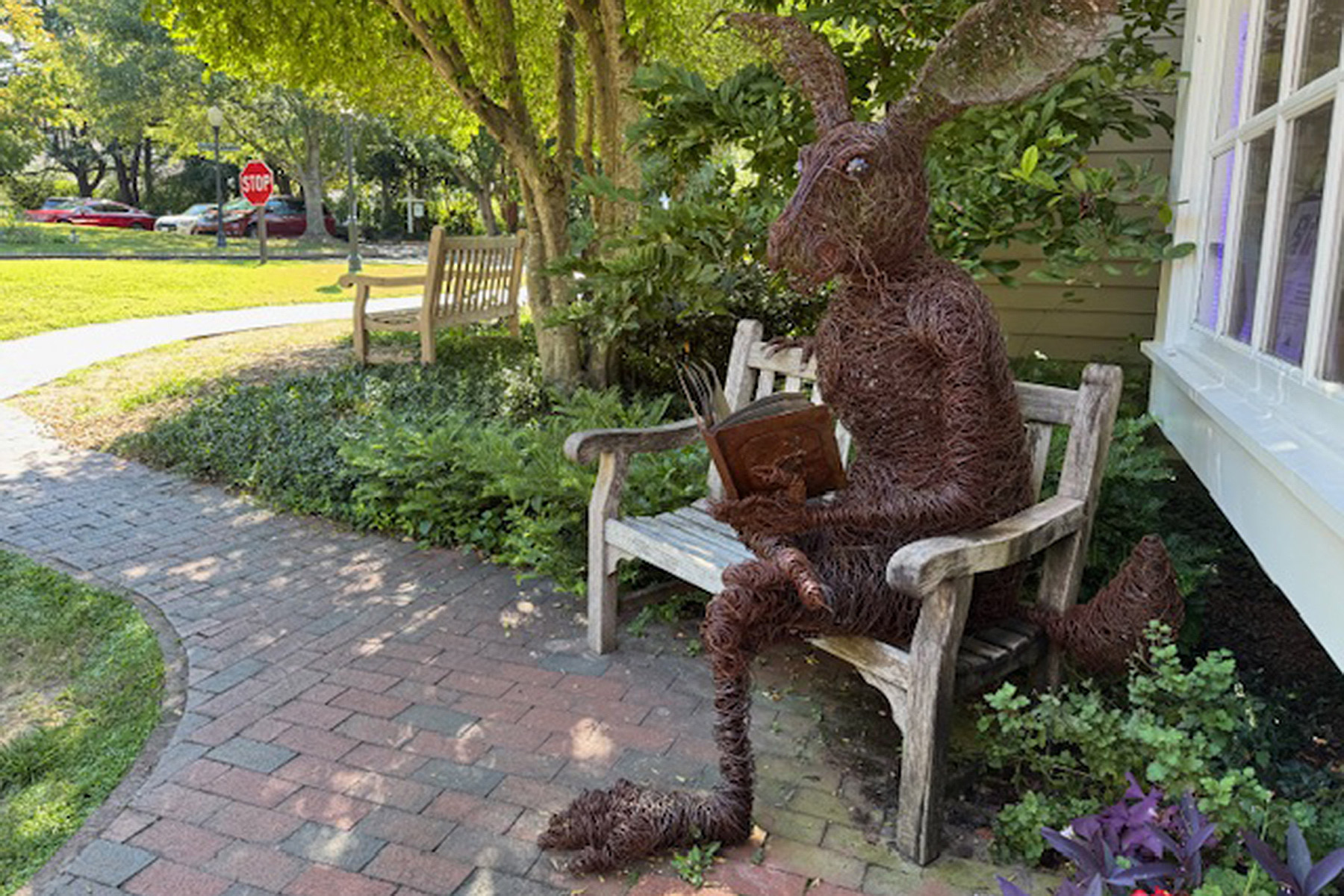 A large wire sculpture of a rabbit sits on a wooden bench, holding a book, in a garden area next to a building with trees, plants, and a brick pathway nearby. Fearrington Village