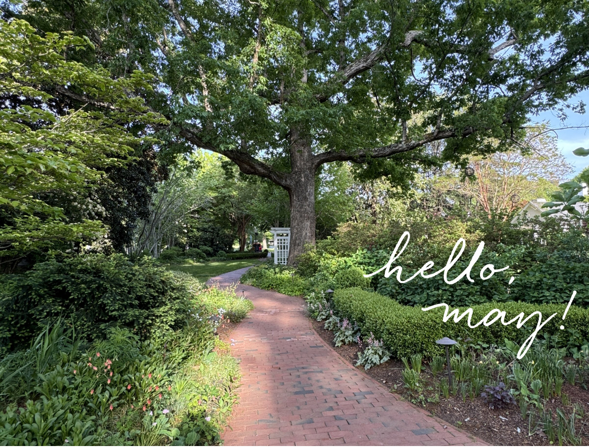 A sunny garden path lined with green trees and flowers curves under a large tree. “Hello, May!” is written in white script on the right side of the image. Fearrington Village