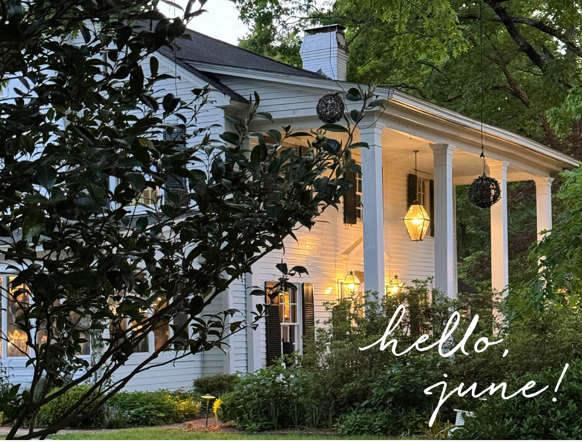 A white house with large columns is surrounded by green trees and bushes at dusk, with warm lights glowing from the porch. The words "hello, june!" are written in white script in the bottom right corner. Fearrington Village