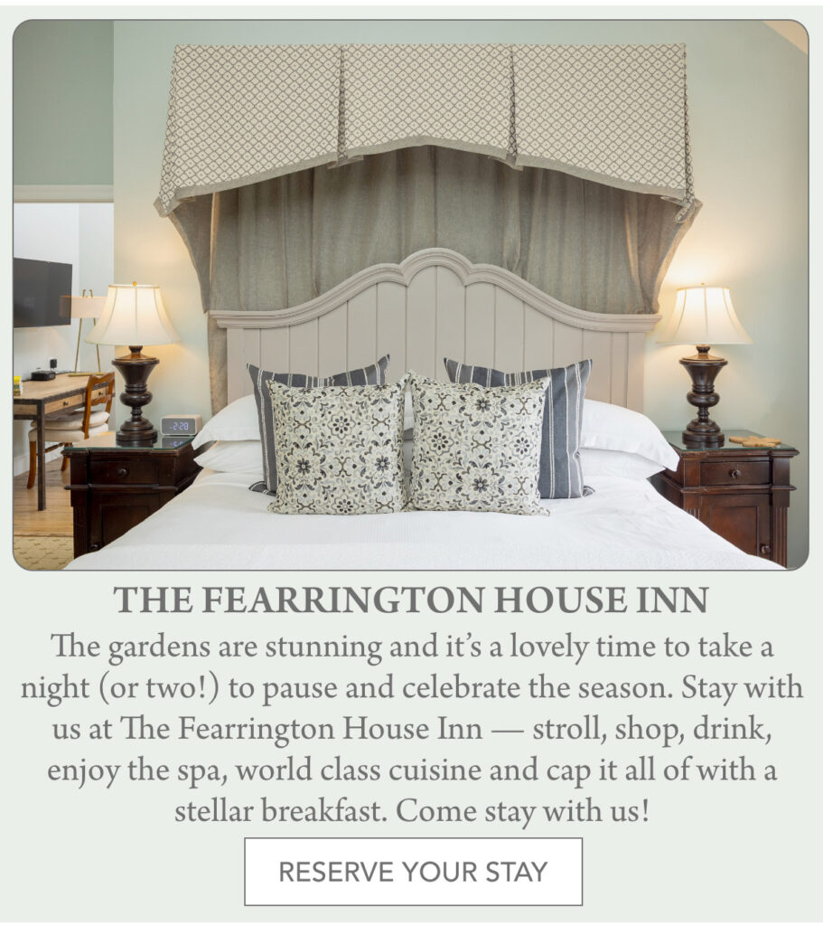 A cozy bedroom at The Fearrington House Inn features a large bed with patterned pillows, matching bedside tables with lamps, and elegant drapery above the headboard. A button reads "RESERVE YOUR STAY." "The gardens are stunning and it’s a lovely time to take a night (or two!) to pause and celebrate the season. Stay with us at The Fearrington House Inn — stroll, shop, drink, enjoy the spa, world class cuisine and cap it all of with a stellar breakfast. Come stay with us!" Fearrington Village