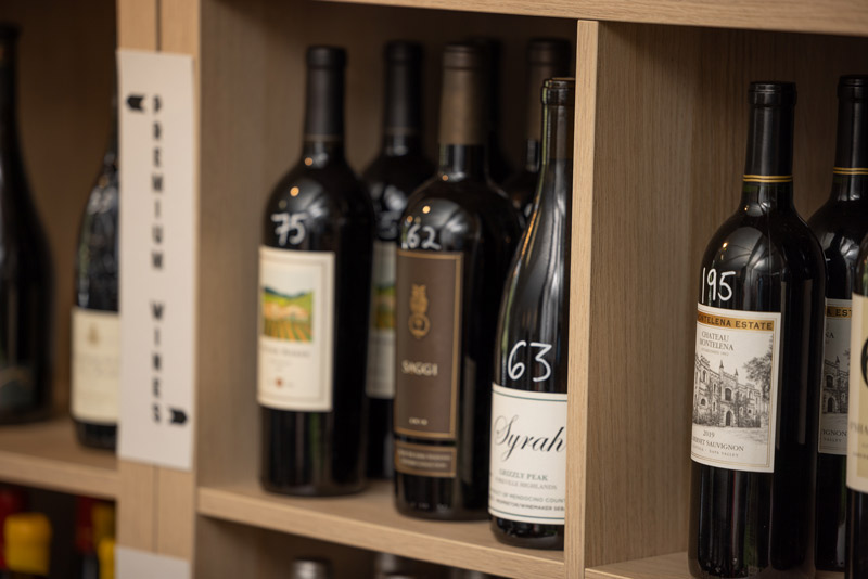 Several bottles of red wine are displayed on wooden shelves. Each bottle has a white number written on it and various label designs, with part of a sign visible on the left. Fearrington Village