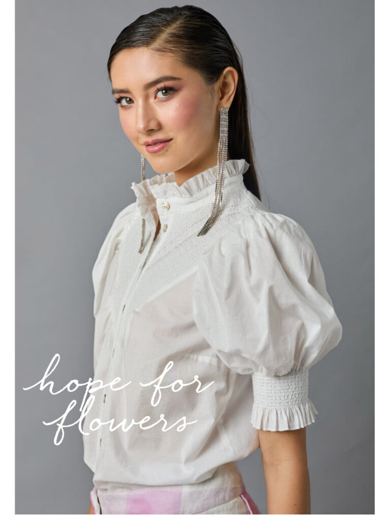 Join us for our Hope for Flowers Trunk Show this Friday & Saturday! 🌿✨ Sustainable. Feminine. 2 A woman in a ruffled white blouse with puffed sleeves and long, sparkling earrings poses against a gray background. White script text on the image reads, "hope for flowers. Fearrington Village