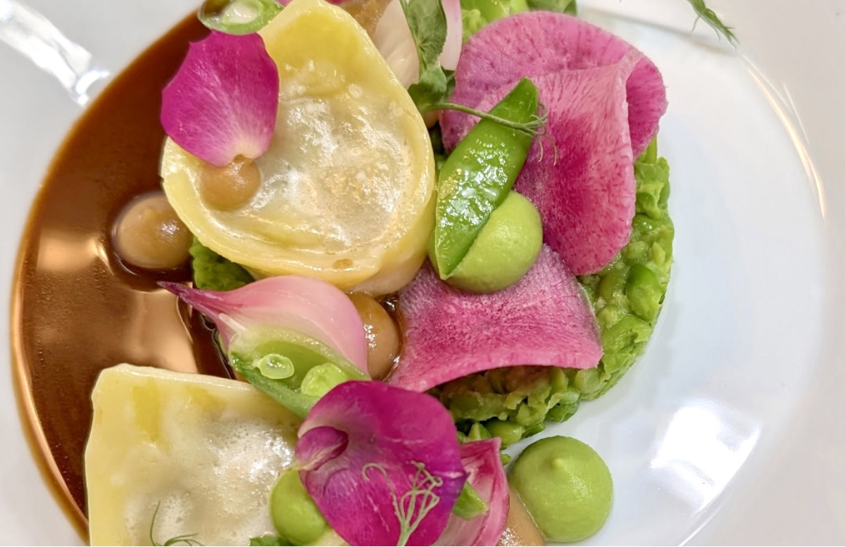 A gourmet dish featuring ravioli, green pea puree, snap peas, pink radish slices, edible flowers, and a brown sauce, artfully arranged on a white plate. Fearrington Village