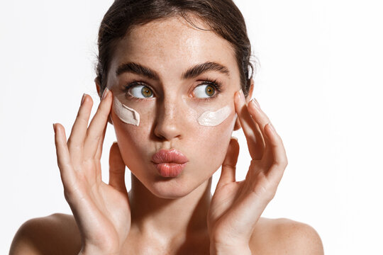 Young woman with brown hair and clear skin applying cream under her eyes, looking to the side with a playful, puckered expression, against a plain white background. Fearrington Village