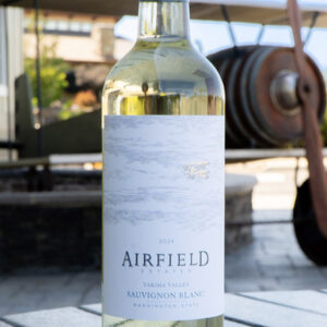 A bottle of Airfield Estates 2024 Sauvignon Blanc wine sits on a stone surface outdoors, with a vintage airplane blurred in the background. Fearrington Village