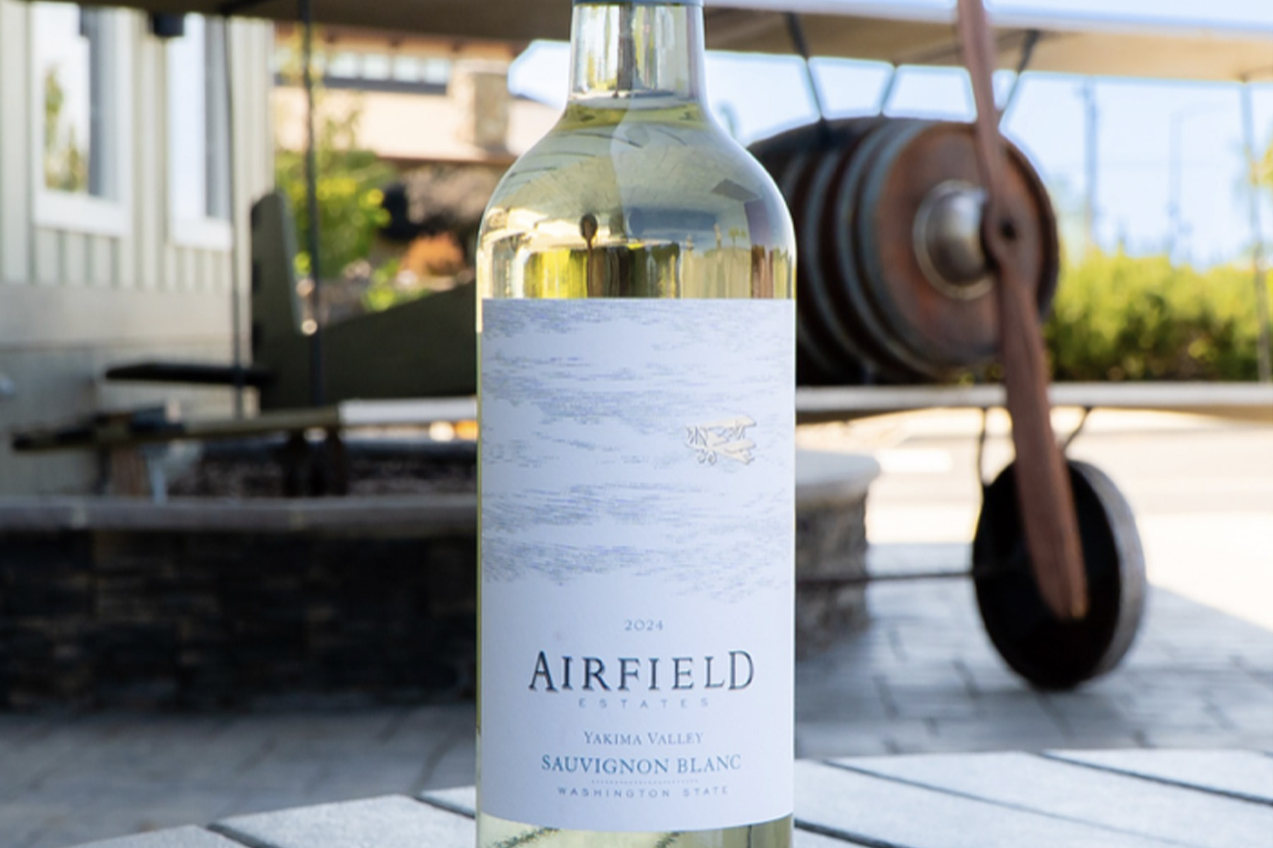 Free Wine Tasting - Airfield 82 A bottle of Airfield Estates 2024 Sauvignon Blanc wine sits on a stone surface outdoors, with a vintage airplane blurred in the background. Fearrington Village