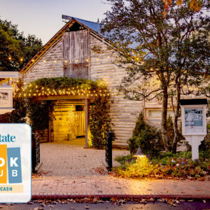 A rustic wooden building with warm string lights glowing around the entrance, surrounded by trees and autumn leaves. In the corner, a "Our State Book Club with Wiley Cash" logo appears. Fearrington Village
