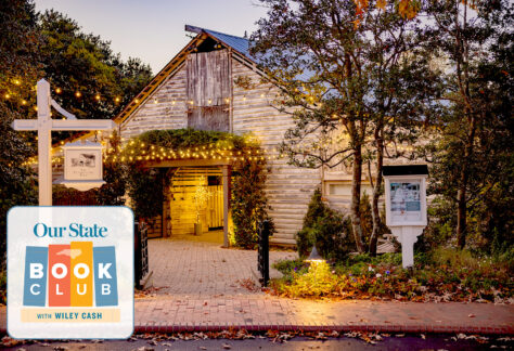 A rustic wooden building with warm string lights glowing around the entrance, surrounded by trees and autumn leaves. In the corner, a "Our State Book Club with Wiley Cash" logo appears. Fearrington Village