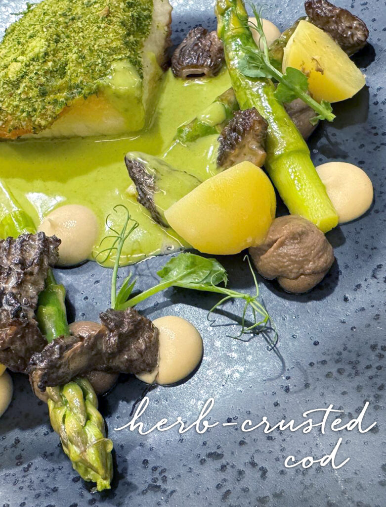 A plated dish of herb-crusted cod with green sauce, asparagus, yellow potatoes, morel mushrooms, and pea shoots, artistically arranged on a dark speckled plate. Text reads "herb-crusted cod. Fearrington Village