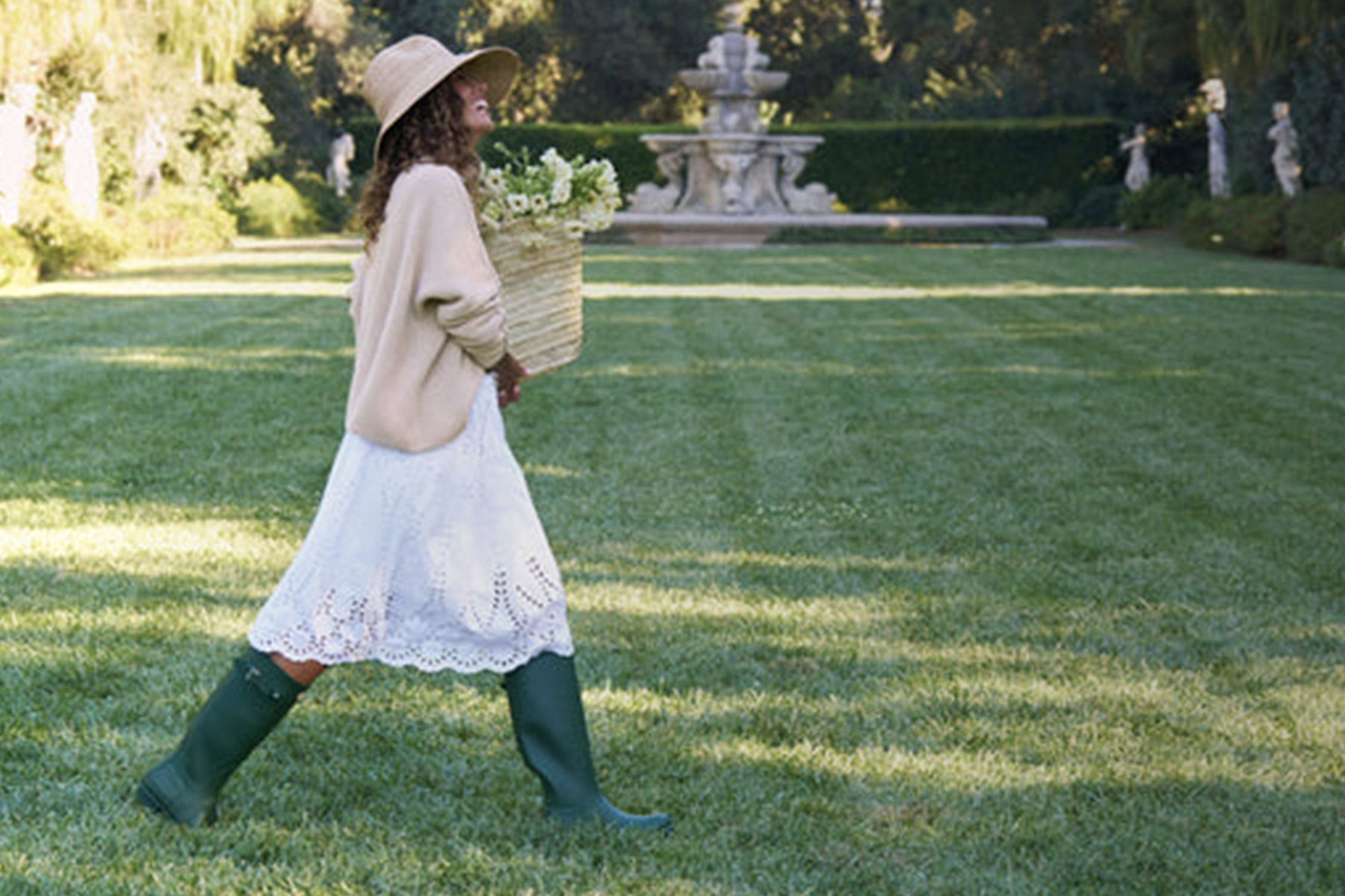 A woman wearing a sun hat, beige sweater, white skirt, and green rain boots walks across a grassy lawn, carrying a woven basket filled with flowers. A fountain and trees are visible in the background. Fearrington Village