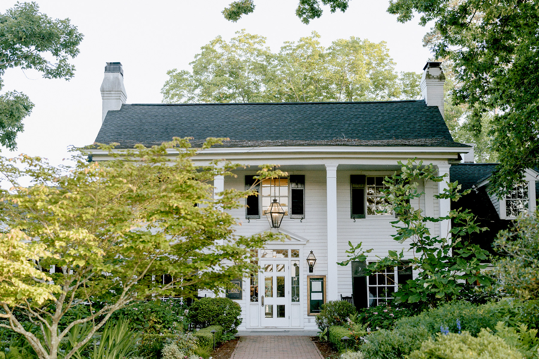 A white two-story house with black shutters, two chimneys, tall columns, and a dark roof, surrounded by lush green trees and plants, under a bright sky. Fearrington Village
