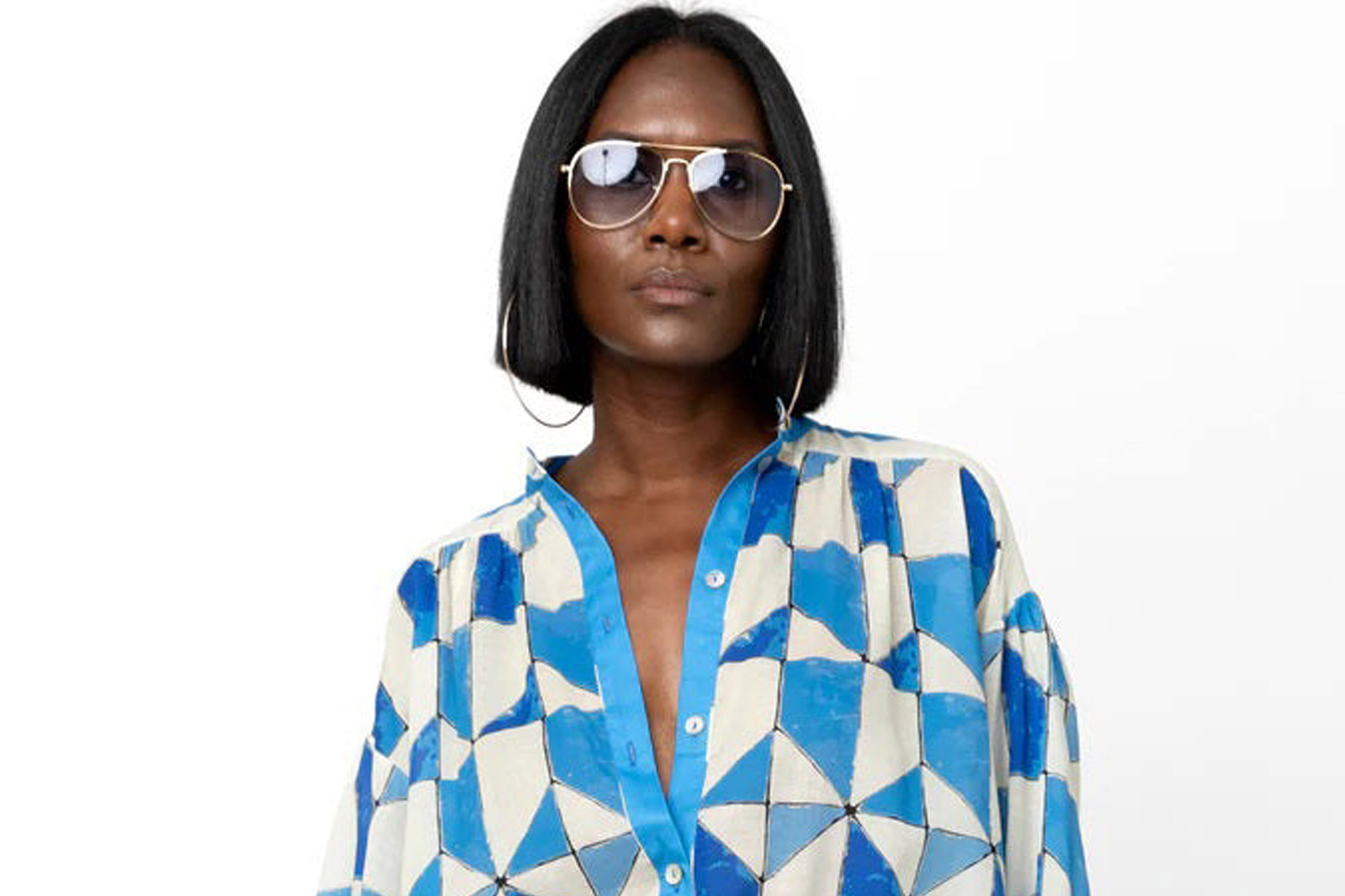 A woman with straight shoulder-length black hair wears sunglasses and a blue-and-white geometric patterned button-up shirt, standing against a plain white background. Fearrington Village