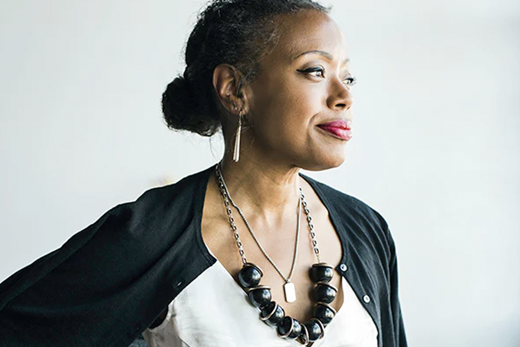 A woman with her hair in a bun gazes to the side. She wears a white top, a black cardigan, a chunky black necklace, and long earrings, with a calm and confident expression against a plain background. Fearrington Village