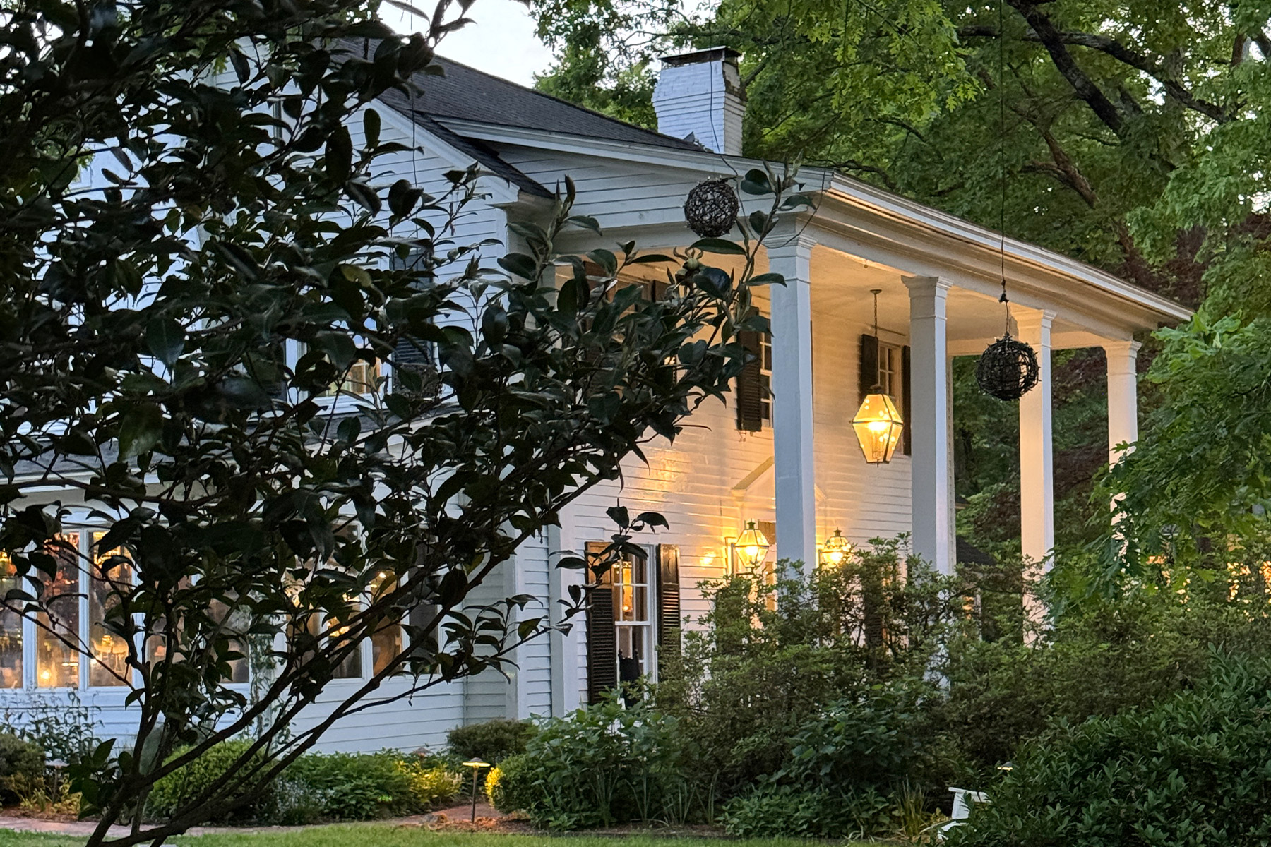 A white house with tall columns is surrounded by lush greenery at dusk. Warm lights glow from inside and outside, creating a cozy atmosphere. A tree partially obscures the view on the left. Fearrington Village