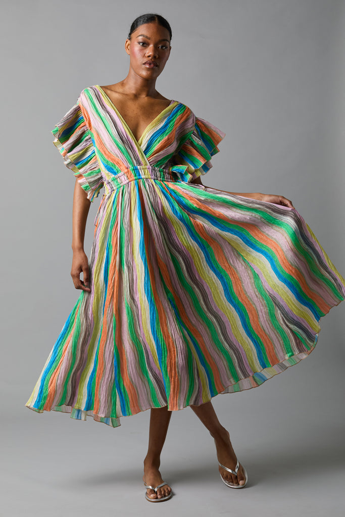 A woman poses in a colorful, knee-length, flowy dress with vertical wavy stripes in shades of green, blue, orange, yellow, and pink. She wears clear sandals and stands against a plain gray background. Fearrington Village