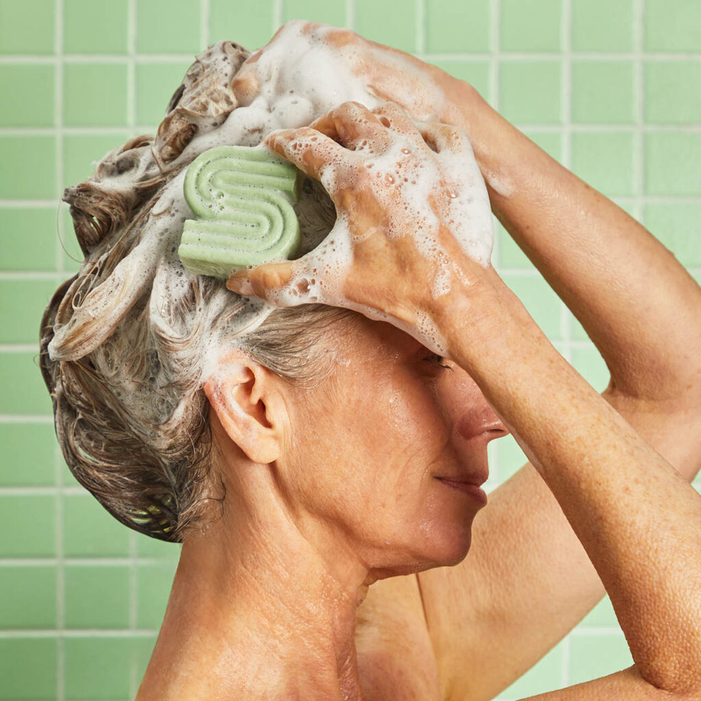 An older woman with gray hair is washing her hair, creating lots of lather with a green shampoo bar in the shape of an "S" against a background of green tiles. Fearrington Village