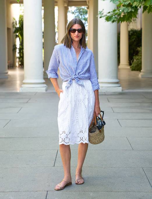 Woman standing outdoors wearing sunglasses, a blue and white striped button-up shirt tied at the waist, a white eyelet skirt, and sandals. She holds a straw bag with dark handles and stands in front of white columns. Fearrington Village
