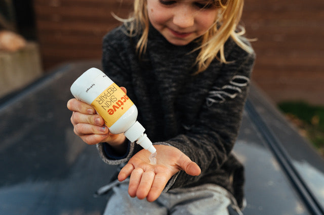 The Spa Edit - First Aid Essentials 1 A young child with long blond hair squirts lotion from a bottle labeled "Active Skin Repair" onto their palm while sitting outdoors. Fearrington Village