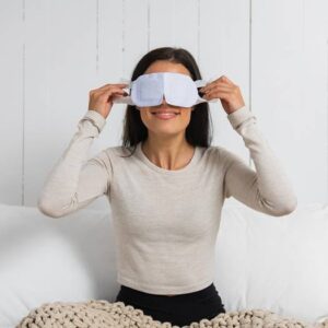 A woman sitting on a bed and smiling while putting on a white eye mask. She is wearing a light long-sleeve shirt and is partially covered with a knitted beige blanket. The background features white wooden panels. Fearrington Village