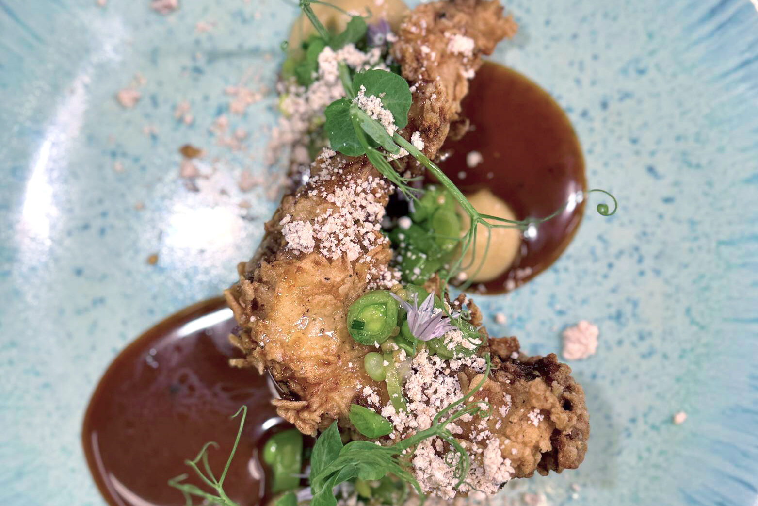The Fearrington Plate - May 15, 2025 1 A gourmet dish on a blue plate featuring crispy fried pieces, vibrant green herbs and peas, creamy dollops, and rich brown sauce artfully arranged with delicate garnish. Fearrington Village