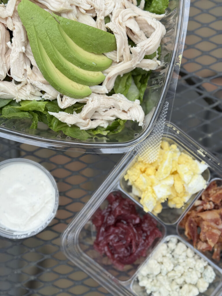 A plastic container holds a salad with shredded chicken, sliced avocado, and lettuce. Next to it are separate compartments with scrambled eggs, bacon, blue cheese, and beets, plus a cup of creamy dressing. Fearrington Village