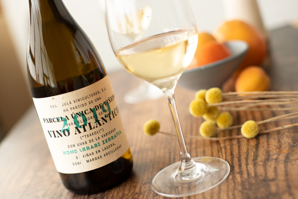 A bottle of white wine and a filled wine glass on a wooden table, with yellow billy button flowers and a blue bowl of oranges blurred in the background. Fearrington Village