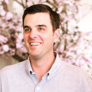 A man with short dark hair, wearing a light gray button-up shirt, smiles while standing in front of a blurred background with pink flowering branches. Fearrington Village