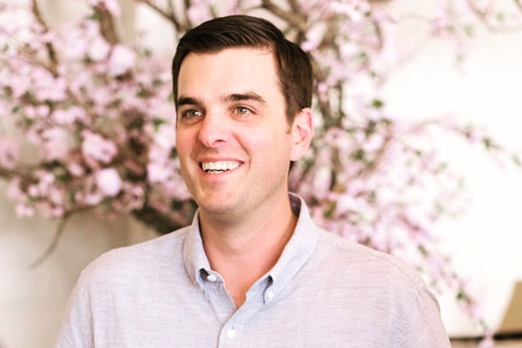 A man with short dark hair, wearing a light gray button-up shirt, smiles while standing in front of a blurred background with pink flowering branches. Fearrington Village
