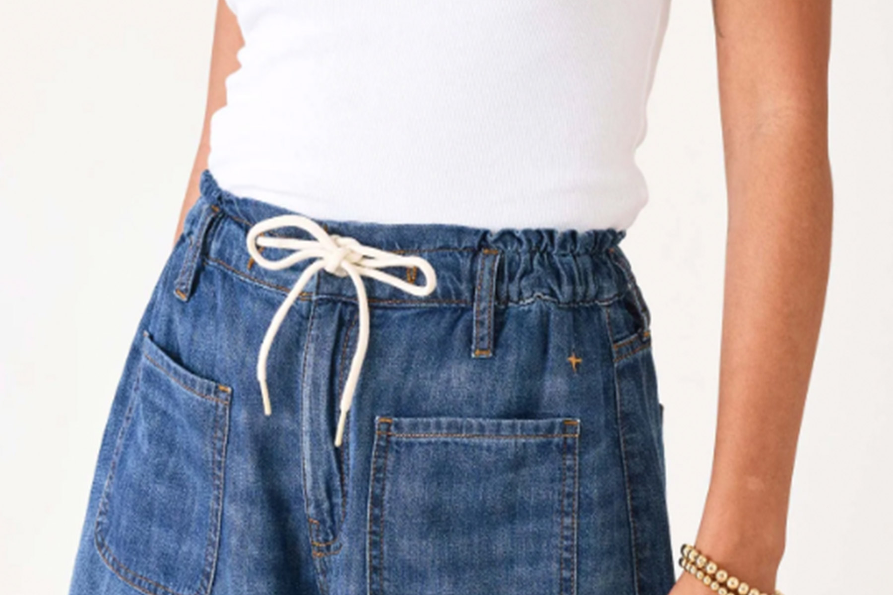 A person wearing a white tank top tucked into high-waisted blue denim pants with an elastic waistband and white drawstring, with one arm visible and a beaded bracelet on their wrist. Fearrington Village