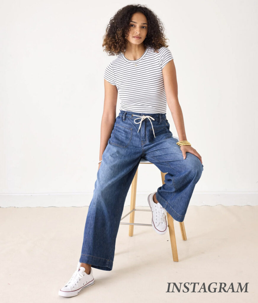 A woman with curly hair, wearing a white and black striped t-shirt, loose blue jeans with a drawstring, and white sneakers, sits on a wooden stool against a plain background. The word "INSTAGRAM" is in the bottom right corner. Fearrington Village