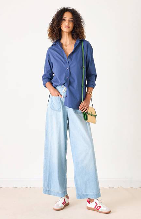 A woman with curly hair wears a blue button-up shirt, light blue wide-leg jeans, white sneakers with red accents, and carries a small green-trimmed crossbody bag. She stands against a plain white background. Fearrington Village
