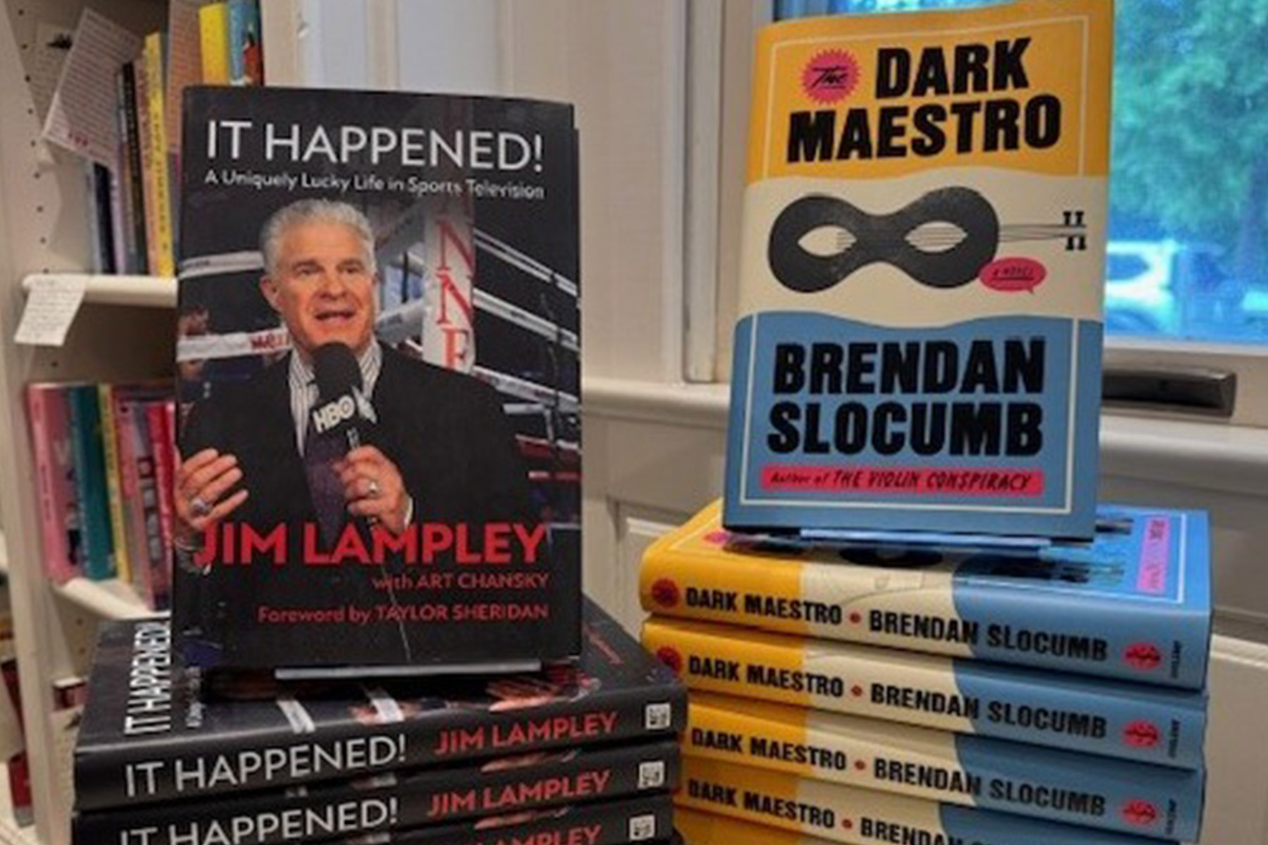 Two books on display: "It Happened!" by Jim Lampley, featuring a man holding a microphone, and "Dark Maestro" by Brendan Slocumb, stacked in a pile with yellow and blue covers. Bookshelves are in the background. Fearrington Village